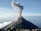 A volcano emits a thick plume of smoke from its summit as it stands prominently against the backdrop of a clear sky. The landscape around the volcano is rugged with scattered rocks in the foreground. White clouds hover gently above the horizon line.
