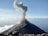 A volcano emits a thick plume of smoke from its summit as it stands prominently against the backdrop of a clear sky. The landscape around the volcano is rugged with scattered rocks in the foreground. White clouds hover gently above the horizon line.