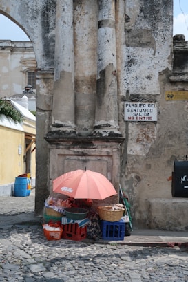 A small street vendor stall is set up against an old, weathered stone wall with protruding columns. The stall includes an umbrella for shade, crates, and baskets filled with goods. A sign on the wall reads 'Parqueo del Santuario Salida No Entre.' The surrounding area features cobblestone pavement and an alley leading to other historical buildings.