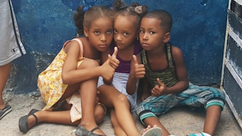 Three children are sitting closely together on the ground in front of a blue wall. Two girls and a boy are seated, with the girls wearing dresses and the boy wearing a tank top and checkered pants. They all have serious expressions and are giving thumbs-up gestures. The setting appears to be an informal outdoor area.