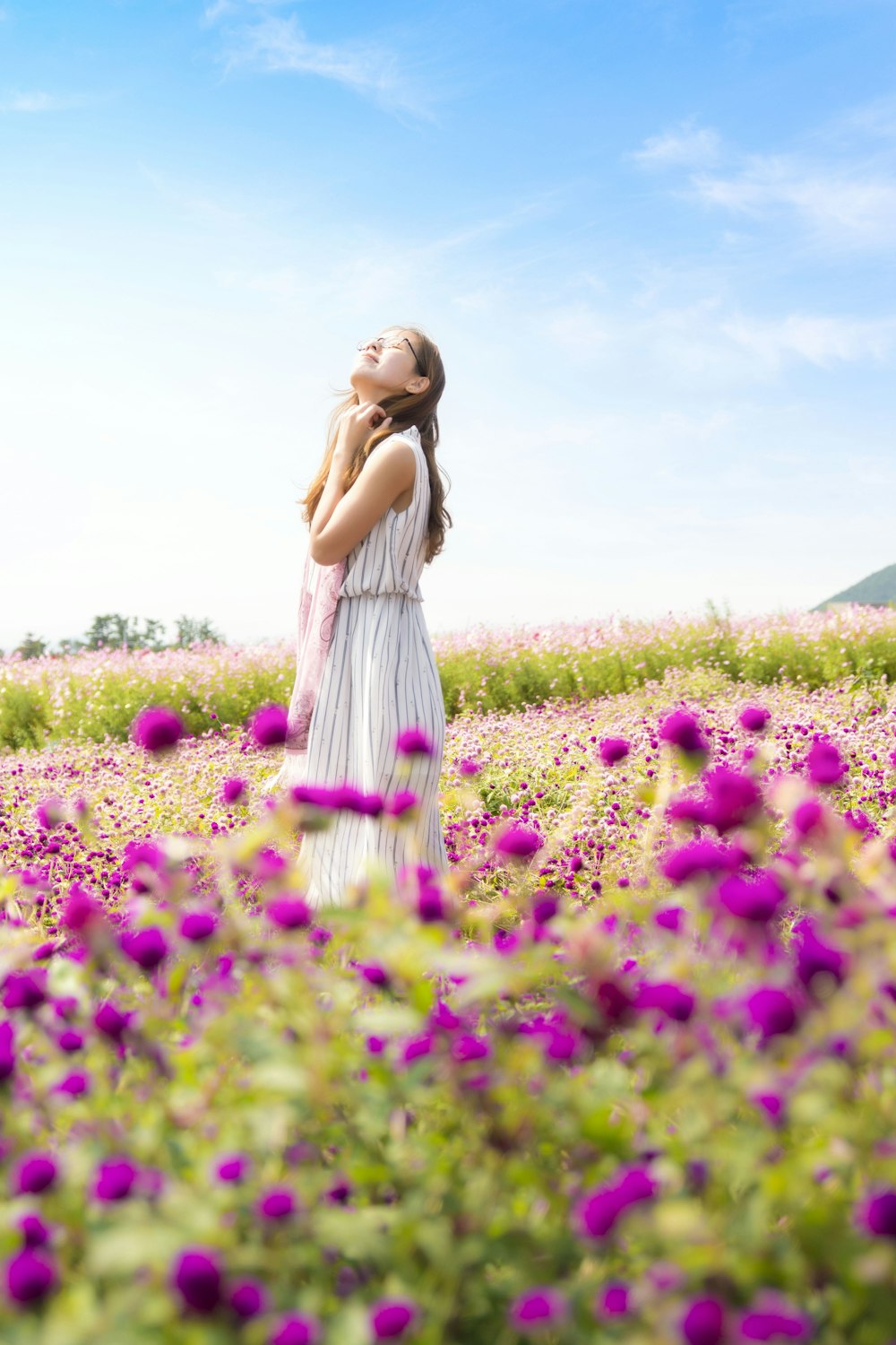 白とグレーの縞模様のノースリーブドレスを着た女性が、昼間のピンクの花畑に立っている空気の匂いを嗅いでいる