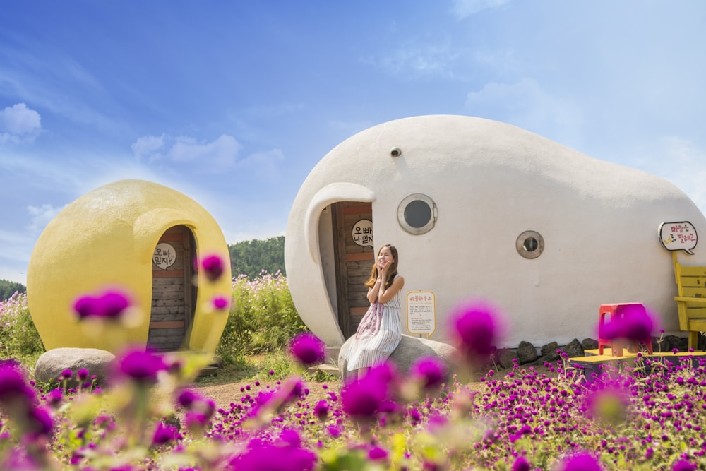 woman sitting in front of white and brown rock house surrounded with purple flowers under blue sky