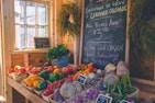 A cozy, rustic market stall displaying a variety of colorful fresh produce such as tomatoes, bell peppers, carrots, and turnips. A blackboard with handwritten notes on organic offerings, pricing, and a chicken advertisement is prominently displayed. Potted herbs and several hanging plants add a touch of greenery to the sunny, wooden interior.