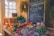 A cozy, rustic market stall displaying a variety of colorful fresh produce such as tomatoes, bell peppers, carrots, and turnips. A blackboard with handwritten notes on organic offerings, pricing, and a chicken advertisement is prominently displayed. Potted herbs and several hanging plants add a touch of greenery to the sunny, wooden interior.
