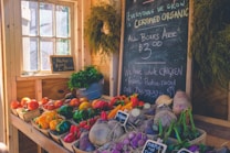 A cozy, rustic market stall displaying a variety of colorful fresh produce such as tomatoes, bell peppers, carrots, and turnips. A blackboard with handwritten notes on organic offerings, pricing, and a chicken advertisement is prominently displayed. Potted herbs and several hanging plants add a touch of greenery to the sunny, wooden interior.