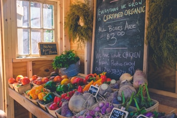 A cozy, rustic market stall displaying a variety of colorful fresh produce such as tomatoes, bell peppers, carrots, and turnips. A blackboard with handwritten notes on organic offerings, pricing, and a chicken advertisement is prominently displayed. Potted herbs and several hanging plants add a touch of greenery to the sunny, wooden interior.
