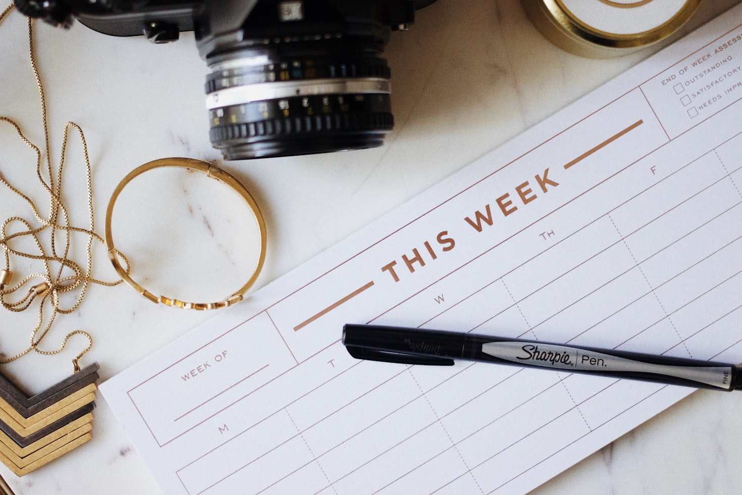 A weekly calendar, sharpie, jewelry, and a camera all sitting on a table top.