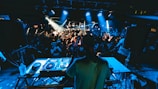 A DJ is performing in front of a lively crowd at a club or concert. The audience is dancing and raising their hands, with bright stage lights illuminating the scene. The DJ booth is equipped with professional sound equipment.
