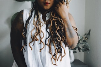 A person with long, multi-colored dreadlocks wearing a sleeveless white shirt. They have tattoos on their arms and are standing indoors next to a table with a white vase containing leaves and branches.