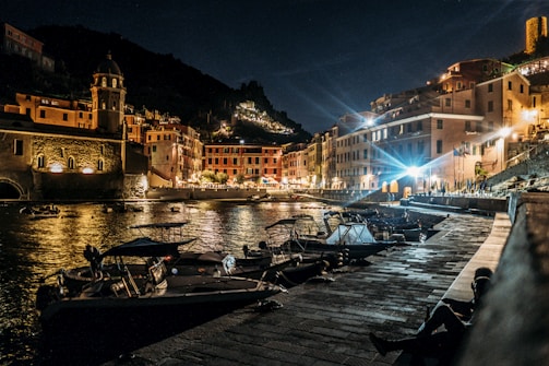 Evening view of a coastal boardwalk with homes lit warmly and boats docked nearby