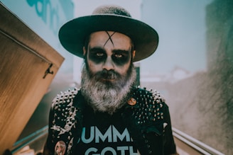 A person with a full beard and dramatic black eye makeup wears a wide-brimmed hat and a studded jacket. Their shirt, partially visible, reads 'GOTH' and the setting appears urban with bright lighting and diffused background.