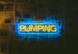 A neon sign displays the word 'PUMPING' in yellow against a blue rectangular background. The sign is mounted on a wall covered with newspaper pages.
