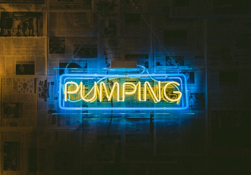 A neon sign displays the word 'PUMPING' in yellow against a blue rectangular background. The sign is mounted on a wall covered with newspaper pages.