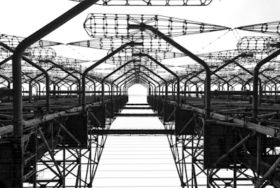 A complex network of metal structures and cables forms an intricate industrial framework. The perspective looks upwards, highlighting the repetitive geometric patterns and intricate lattice work.