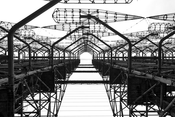 A complex network of metal structures and cables forms an intricate industrial framework. The perspective looks upwards, highlighting the repetitive geometric patterns and intricate lattice work.