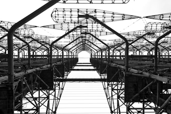 A complex network of metal structures and cables forms an intricate industrial framework. The perspective looks upwards, highlighting the repetitive geometric patterns and intricate lattice work.