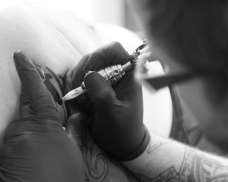 A tattoo artist is applying a tattoo on a person's skin using a tattoo machine. The artist is wearing black gloves and holding the machine with precision. The tattoo appears to have bold black lines.