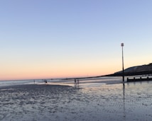 A serene beach scene during low tide with a wide expanse of wet sand reflecting the clear sky above. The horizon is painted in pastel shades of pink and blue as the sun sets. A few people can be seen walking along the water's edge, and a navigational marker stands tall on the right side near the shore. Distant hills and a series of wooden groynes stretch towards the sea.