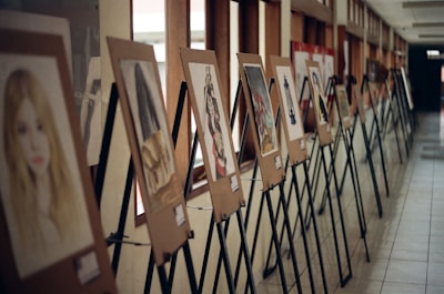 A hallway display of artworks mounted on wooden easels, with a series of painted portraits and abstract images arranged in a line. The display is set against a wall with large windows, allowing natural light to illuminate the pieces.