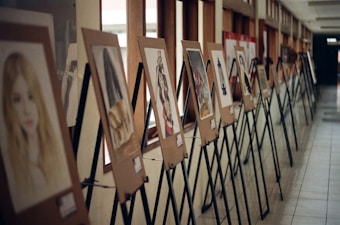 A hallway display of artworks mounted on wooden easels, with a series of painted portraits and abstract images arranged in a line. The display is set against a wall with large windows, allowing natural light to illuminate the pieces.
