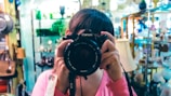 A person wearing a pink shirt holds up a Canon camera, obscuring their face while taking a photograph. They are in a store or showroom filled with various items such as lamps, bags, and decorative objects. The background displays shelves with glassware and other merchandise, giving the setting a cozy, vintage vibe.