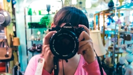 A person wearing a pink shirt holds up a Canon camera, obscuring their face while taking a photograph. They are in a store or showroom filled with various items such as lamps, bags, and decorative objects. The background displays shelves with glassware and other merchandise, giving the setting a cozy, vintage vibe.