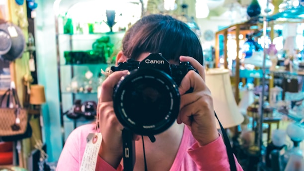 A person wearing a pink shirt holds up a Canon camera, obscuring their face while taking a photograph. They are in a store or showroom filled with various items such as lamps, bags, and decorative objects. The background displays shelves with glassware and other merchandise, giving the setting a cozy, vintage vibe.