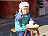 A child wearing a colorful outfit, including a bright blue and green puffy jacket and a whimsical animal-shaped hat, is sitting on a wooden stool outside. The child is eating from a cup using a spoon, appearing contemplative and focused.