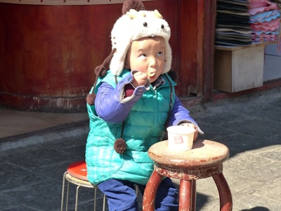 A child wearing a colorful outfit, including a bright blue and green puffy jacket and a whimsical animal-shaped hat, is sitting on a wooden stool outside. The child is eating from a cup using a spoon, appearing contemplative and focused.