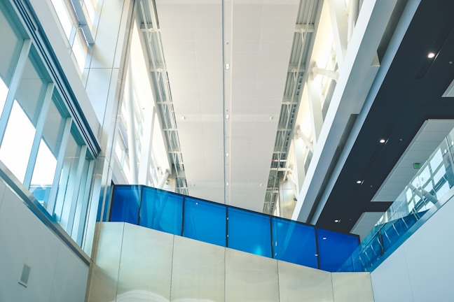 A modern architectural interior featuring high ceilings, large windows allowing abundant natural light, and sleek design elements. The use of glass and metal provides a contemporary feel, with a prominent blue glass panel adding a splash of color.