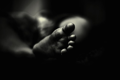 A black and white photograph features the close-up of a foot, focusing on the toes and capturing intricate details. The foot is softly lit, creating a contrast between light and shadow that adds depth to the image. The background fades into darkness, emphasizing the foreground subject.