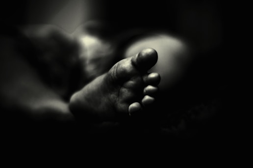 A black and white photograph features the close-up of a foot, focusing on the toes and capturing intricate details. The foot is softly lit, creating a contrast between light and shadow that adds depth to the image. The background fades into darkness, emphasizing the foreground subject.