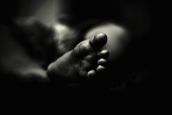 A black and white photograph features the close-up of a foot, focusing on the toes and capturing intricate details. The foot is softly lit, creating a contrast between light and shadow that adds depth to the image. The background fades into darkness, emphasizing the foreground subject.