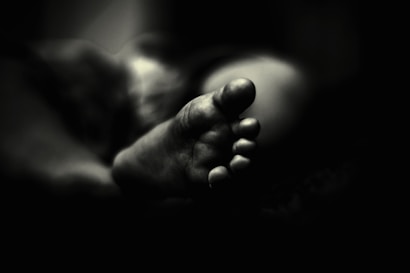 A black and white photograph features the close-up of a foot, focusing on the toes and capturing intricate details. The foot is softly lit, creating a contrast between light and shadow that adds depth to the image. The background fades into darkness, emphasizing the foreground subject.