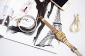 An open magazine features fashion illustrations and perfume advertisements with elegant typography. A silver and gold bracelet lies over the page, along with a gold wristwatch displaying the time. A smartphone and a small perfume bottle are also placed nearby, creating a stylish and sophisticated aesthetic.