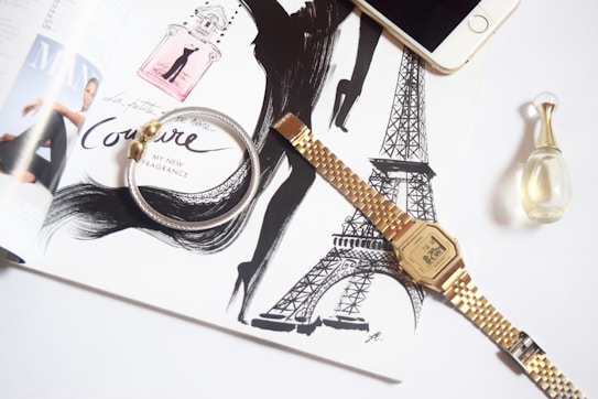 An open magazine features fashion illustrations and perfume advertisements with elegant typography. A silver and gold bracelet lies over the page, along with a gold wristwatch displaying the time. A smartphone and a small perfume bottle are also placed nearby, creating a stylish and sophisticated aesthetic.