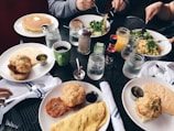 A variety of freshly prepared breakfast items including pancakes with butter, an omelette, biscuits, salad, and crispy fried food are spread across a table with several mugs and mason jars filled with drinks. There are condiments like syrup, coffee, and jam. The setting includes people holding utensils and eating.