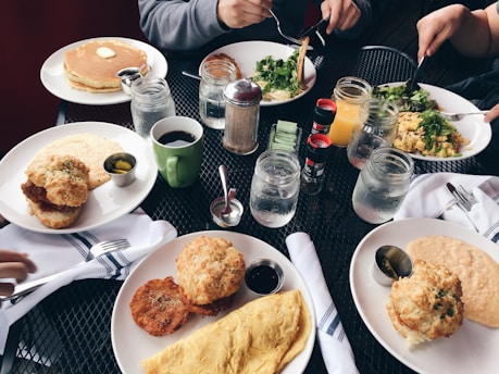 A variety of freshly prepared breakfast items including pancakes with butter, an omelette, biscuits, salad, and crispy fried food are spread across a table with several mugs and mason jars filled with drinks. There are condiments like syrup, coffee, and jam. The setting includes people holding utensils and eating.