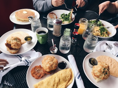 A variety of freshly prepared breakfast items including pancakes with butter, an omelette, biscuits, salad, and crispy fried food are spread across a table with several mugs and mason jars filled with drinks. There are condiments like syrup, coffee, and jam. The setting includes people holding utensils and eating.