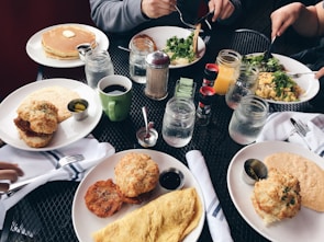 A variety of freshly prepared breakfast items including pancakes with butter, an omelette, biscuits, salad, and crispy fried food are spread across a table with several mugs and mason jars filled with drinks. There are condiments like syrup, coffee, and jam. The setting includes people holding utensils and eating.