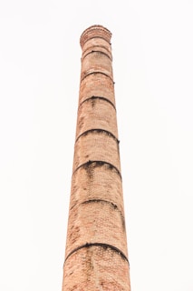 A tall, cylindrical brick chimney extends vertically against a light, overcast sky. The structure features a series of black metal bands at regular intervals and a textured surface with visible brickwork.