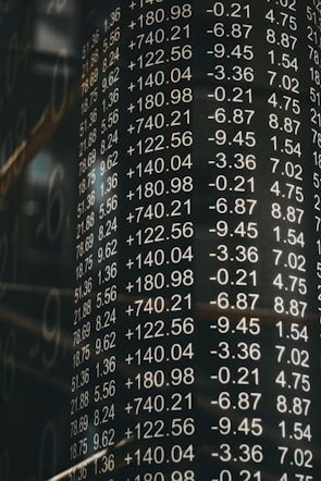 A board displays various financial numbers, including positive and negative values with decimal points. The numbers are aligned in columns, suggesting a stock market or financial data context. The overall scene conveys a sense of an analytic or financial environment.