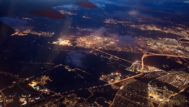 Aerial view of a city at night with a network of illuminated roads and streets. City lights create a glowing web-like pattern against the dark landscape. Clouds are partially visible, and an airplane wing is in the upper part of the image.