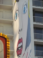 A large pop art mural on the side of a building depicts a dramatic face with wide blue eyes and red lips. The building features balconies in the background, and there is a partially visible colorful sign with yellow circular lights in the foreground.