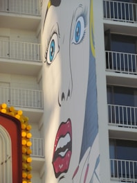 A large pop art mural on the side of a building depicts a dramatic face with wide blue eyes and red lips. The building features balconies in the background, and there is a partially visible colorful sign with yellow circular lights in the foreground.