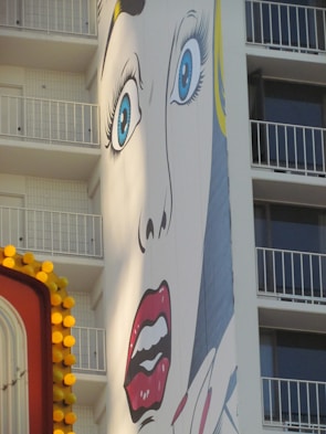 A large pop art mural on the side of a building depicts a dramatic face with wide blue eyes and red lips. The building features balconies in the background, and there is a partially visible colorful sign with yellow circular lights in the foreground.
