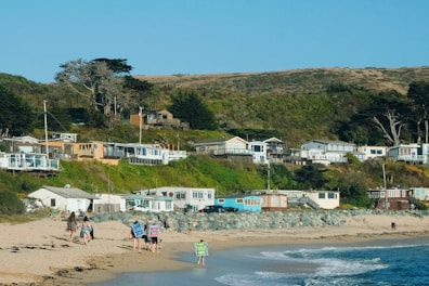 A coastal village with several small houses situated on a grassy hillside near the ocean. People are walking along the sandy beach, some carrying items like backpacks and beach gear. The landscape features a mix of greenery, including trees and shrubs.