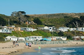 A coastal village with several small houses situated on a grassy hillside near the ocean. People are walking along the sandy beach, some carrying items like backpacks and beach gear. The landscape features a mix of greenery, including trees and shrubs.