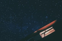 A starry night sky with countless tiny, sparkling stars spread across a dark blue canvas. In the foreground, the corner of a building with an illuminated window is visible, casting a warm orange glow, creating contrast with the cool tones of the night sky.