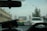 The scene is from inside a vehicle, looking through the front windshield at traffic on a bridge. Cars are lined up, with a green and yellow taxi in front. The bridge structure is visible ahead with towers and overhead traffic lights. To the right, there is a large tree and to the left, a row of buildings with satellite dishes and utility wires.